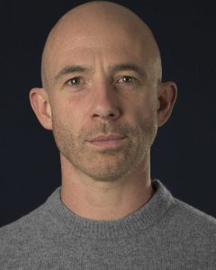 Kevin Marron wearing gray sweater on dark background.